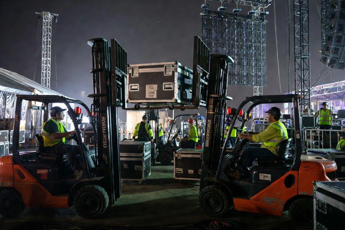 Two forklifts working nose-to-nose at night, a road case raised between them, hi-vis crew moving through the gap.