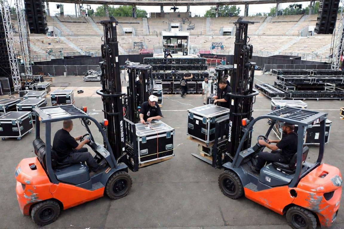 Two counterbalance forklifts facing each other across an amphitheater floor, each carrying a pallet-mounted road case, operators seated with hands on the controls.