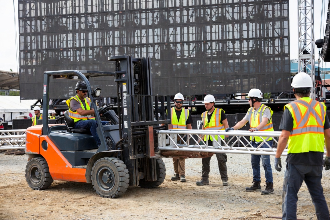 Four crew members walk a length of truss alongside a moving forklift, hands on the load, in front of an LED wall.
