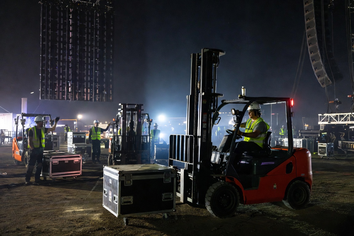 Night festival floor with a large LED wall flown overhead, a forklift positioning a case as a spotter signals.