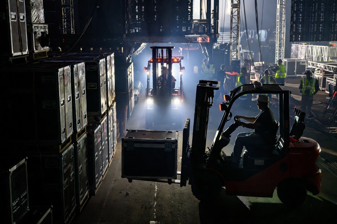 Near-dark load-out: a forklift silhouetted in an alley of stacked road cases, headlights of a second machine glaring head-on through haze.