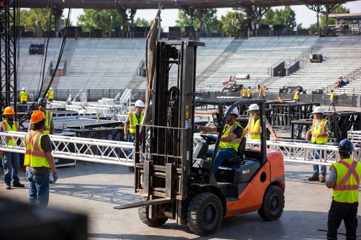 Wide amphitheater load-in with a forklift moving through a large hi-vis crew, truss sections staged across the deck.
