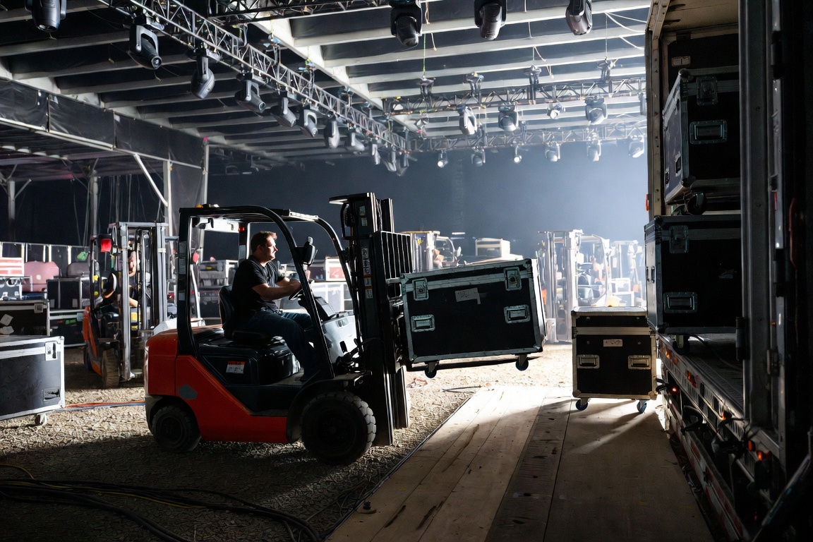 A forklift carries a road case up a timber ramp into the back of a trailer, lighting truss overhead.